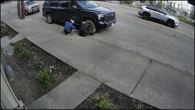 Vigil S4 High alert — two people crouched at front wheel of parked pickup on public sidewalk, May 2026